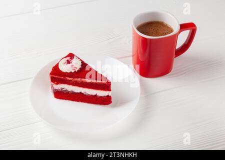 Ein Stück roter Samtkuchen mit cremeweißer Zuckerguss auf einem Holztisch, begleitet von einer dampfenden Tasse aromatischem Kaffee. Stockfoto