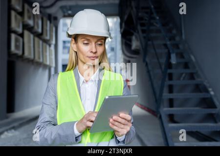 Ernsthaft selbstbewusst denkende Ingenieurin mit Tablet-Computer, der die Fabrik mit Schutzhelm und reflektierender Weste inspiziert. Stockfoto