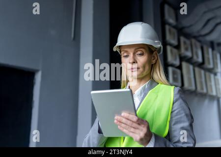 Ernsthaft selbstbewusst denkende Ingenieurin mit Tablet-Computer, der die Fabrik mit Schutzhelm und reflektierender Weste inspiziert. Stockfoto