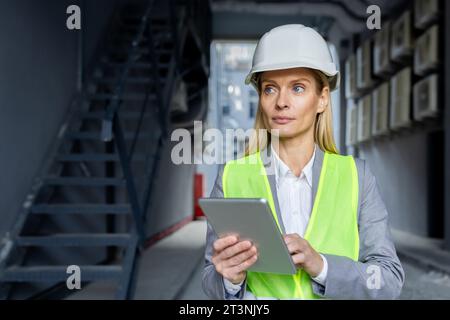 Ernsthaft selbstbewusst denkende Ingenieurin mit Tablet-Computer, der die Fabrik mit Schutzhelm und reflektierender Weste inspiziert. Stockfoto