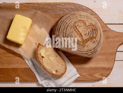 Flacher, glutenfreier Sauerteig mit Herzschlag, aufgeschnitten und gebuttert, zusammen mit einem Butterblock auf einer Holzbrotplatte. Stockfoto
