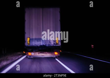 Eine verschwommene Bewegungsaufnahme eines Lkws, der nachts auf einer Autobahn fährt, wobei die Rückleuchten und das Heck des Lkws vor dunkler Kulisse hervorgehoben werden Stockfoto