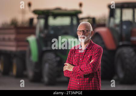 Porträt eines Seniorenbauers, der vor zwei Traktoren mit Anhängern zur Erntezeit steht Stockfoto