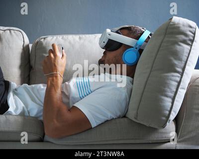 Mann, der ein vr-Headset der neuen Generation zur Unterhaltung trägt und auf dem Sofa liegt. Stockfoto