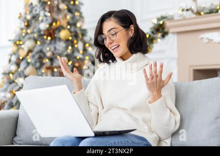 Eine junge Frau sitzt zu Hause auf dem Sofa an Silvesterferien in der Nähe der Badewanne und spricht online auf einem Laptop. Gesten mit Händen, Lächeln. Stockfoto