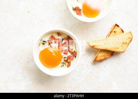 Gebackene Eier (Eggs en cocotte) in Romekins und geröstetes Brot zum Tauchen über hellem Steinhintergrund. Draufsicht, flach Stockfoto