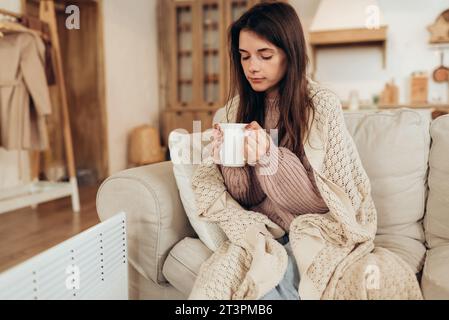 Junge Frau in warmen Kleidern und Karo, die eine Tasse heißes Getränk hält, während sie zu Hause auf der Couch in der Nähe der elektrischen Heizung sitzt. Hochwertige Fotos Stockfoto