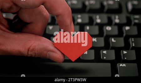 Ein Mann, der ein rotes Origami-Papierherz in der Hand hält über einer schwarzen Computertastatur. Online-Dating-Konzept. Stockfoto