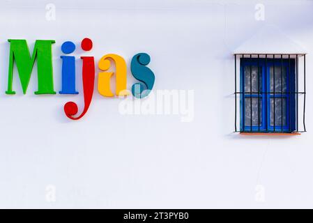 Weiße Wand mit blauem Fenster und dem mehrfarbigen Mijas-Logo. Mijas Pueblo. Mijas, Málaga, Andalusien, Spanien, Europa Stockfoto