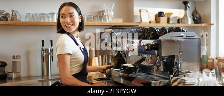 Ein lächelndes asiatisches Barista-Mädchen macht Cappuccino mit einer Kaffeemaschine, steht hinter der Theke im Café Stockfoto