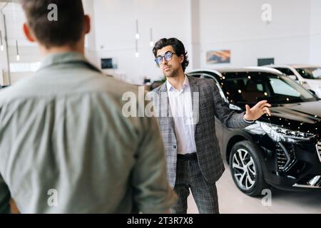 Professioneller Autoverkäufer im Business-Anzug, der nicht erkennbaren interessierten männlichen Käufer über ein luxuriöses Auto in der Autoshow erzählt. Rückansicht des Kunden Stockfoto