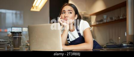 Porträt einer koreanischen Frau, Barista im Café, sah traurig und runzelig aus, grimmige Enttäuschung während der Arbeit im Café, lehnte sich an die Theke, stand Stockfoto