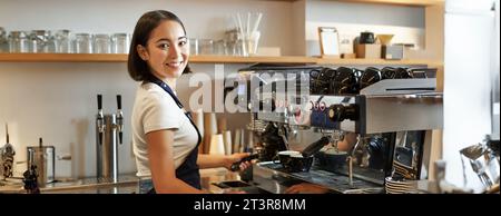 Ein lächelndes asiatisches Barista-Mädchen macht Cappuccino mit einer Kaffeemaschine, steht hinter der Theke im Café Stockfoto