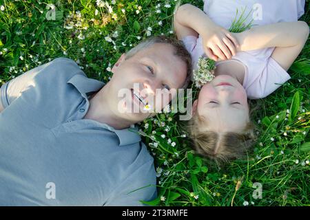 Vater und Tochter liegen auf der Wiese mit Gänseblümchen. Das Mädchen hält einen Strauß Gänseblümchen in der Hand. Weiße Menschen, dunkle Blondinen. Der Mann trägt ein graues T-Shirt, Kind Stockfoto