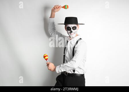 Junger Mann mit bemaltem Schädel und Maracas auf hellem Hintergrund. Mexikanischer Tag der Toten (El Dia de Muertos) Stockfoto