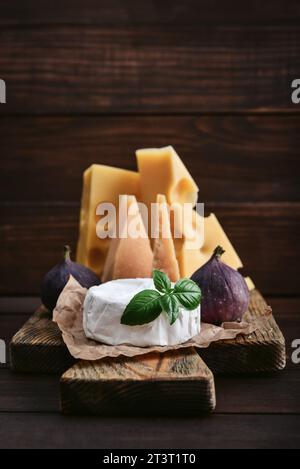 Verschiedene Käsesorten: Camembert, Maasdam, Parmesan - auf einem Schneidebrett mit frischem Basilikum und Feigen auf hölzernem Hintergrund Stockfoto