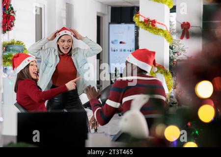 Kollegen haben Spaß in festlich dekorierten Büros und machen sich während der Winterferien gegenseitig auf der Arbeit lustig. Mitarbeiter lachen zusammen in weihnachtlich verzierten Arbeitsbereichen Stockfoto