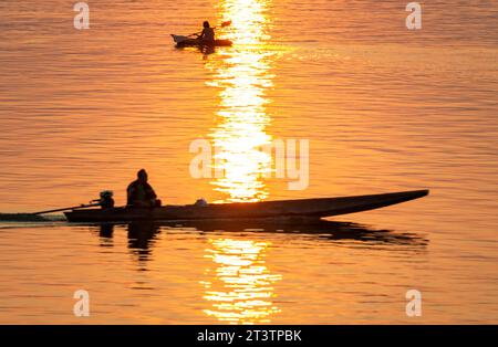 Silhouetten einer menschlichen Gestalt in kleinen Booten, die durch goldene Lichtstrahlen auf dem Wasser über das ruhige, friedliche Wasser des Mekong gleiten Stockfoto