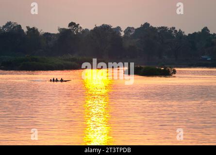 Silhouetten menschlicher Figuren in einem kleinen Kanu, die durch goldene Lichtstrahlen auf dem Wasser über das ruhige, friedliche Wasser des Mekong gleiten Stockfoto