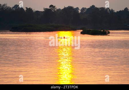 Silhouetten menschlicher Figuren in einem kleinen Kanu, die durch goldene Lichtstrahlen auf dem Wasser über das ruhige, friedliche Wasser des Mekong gleiten Stockfoto