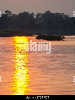 Silhouetten menschlicher Figuren in einem kleinen Kanu, die durch goldene Lichtstrahlen auf dem Wasser über das ruhige, friedliche Wasser des Mekong gleiten Stockfoto