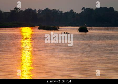 Silhouetten menschlicher Figuren in einem kleinen Kanu, die durch goldene Lichtstrahlen auf dem Wasser über das ruhige, friedliche Wasser des Mekong gleiten Stockfoto