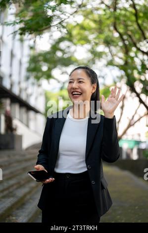 Eine fröhliche und charmante Millennials asiatische Geschäftsfrau in einem formellen Business-Anzug winkt mit der Hand, um ihre Kollegen zu begrüßen, während sie draußen spazieren geht. Stockfoto