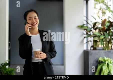 Eine attraktive und selbstbewusste Millennials asiatische Geschäftsfrau ruft jemanden an oder spricht mit jemandem am Telefon, während sie in ihrem Büro läuft. Busine Stockfoto