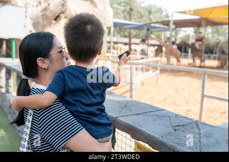 Der zweijährige Junge und seine Mutter beobachten Giraffen in einem Zoo im Freien. Sie stehen neben einem Käfig mit Tieren, Mutter hält einen Jungen im Arm Stockfoto