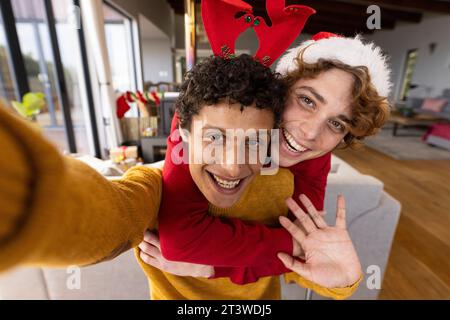 Porträt eines glücklichen, vielseitigen schwulen Paares, das weihnachtsmützen trägt und zu Hause Videoanrufe macht Stockfoto