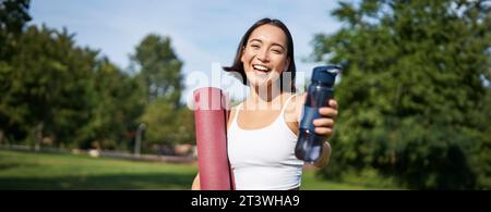 Porträt eines glücklichen asiatischen Mädchens, eine Fitnessfrau, die dir nach dem Training eine Flasche Wasser zum Trinken gibt, mit einer Gummimatte für Übungen im Park Stockfoto