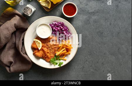 Schnitzel mit Kartoffelfritten, Rotkohl-Salat und Soße auf weißer Platte über dunklem Steinhintergrund mit Kopierraum. Draufsicht, flach Stockfoto