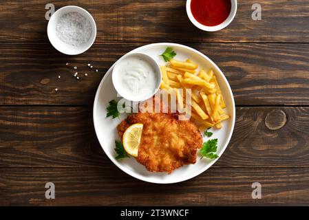 Paniertes Weiner Schnitzel mit Kartoffelfritten und Soße auf weißem Teller auf hölzernem Hintergrund. Draufsicht, flach Stockfoto