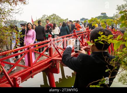 Eine Vietnamese mittleren Alters, die einen rosa ao dai trägt, lässt ihr Mann auf der Huc-Brücke, die zum Ngoc-Son-Tempel am Hoan-Kiem-See führt, fotografieren Stockfoto