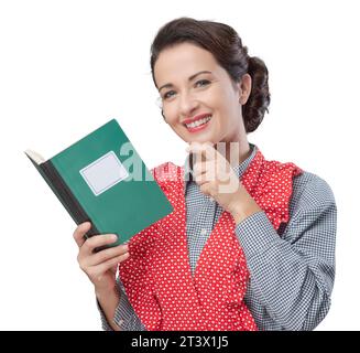 Lächelnde Retro-Frau in Schürze mit Kochbuch und Hand am Kinn Stockfoto