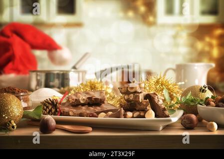 Stücke hausgemachte Schokolade mit Haselnüssen auf weißer Keramikplatte auf hölzerner Küchenbank mit Zutaten und Küchenutensilien und Background-Küche Stockfoto