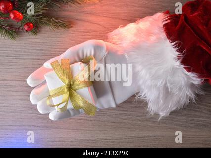 Weihnachtsmann mit weißer Geschenkbox in der Hand mit goldener Schleife auf Holztisch mit bunten Lichtern und Tannenzweigen. Draufsicht. Stockfoto
