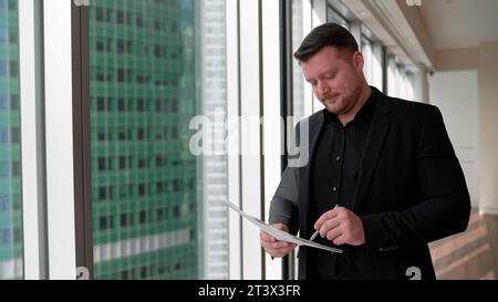 Geschäftsmann mit Klemmbrett, der vor dem neuen Gebäude steht. Ein Geschäftsmann in einem Anzug unterschreibt Dokumente vor dem Hintergrund von Wolkenkratzern. Stockfoto