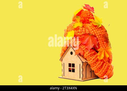Holzspielhaus mit einem klobigen Pompon Hut, großzügig mit Herbstblättern bestreut auf gelbem Hintergrund mit Kopierraum. Stockfoto