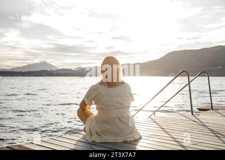 Eine Frau in gelbem Kleid sitzt auf einem Damm am See bei Sonnenuntergang Stockfoto