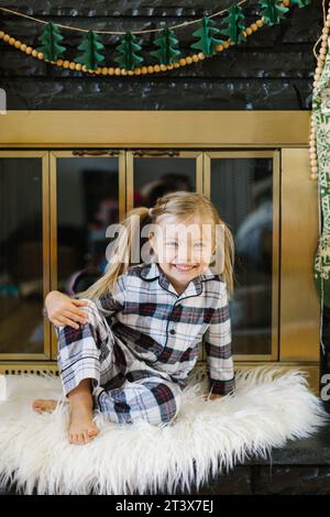 Fröhliches Mädchen (3) im karierten Pyjama sitzt auf Teppich am Kamin Stockfoto