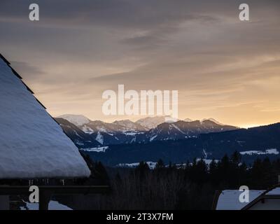 Farbenfroher Winterschneeuntergang über einem österreichischen alpendorf mit Bergen im Hintergrund, schneebedeckten Dächern, Kärnten, Österreich Stockfoto