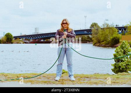 Das Langporträt einer selbstbewussten jungen blonden Frau, die Gartenschlauch hält Stockfoto