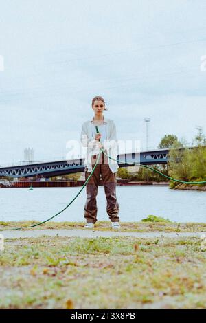 Porträt eines selbstbewussten jungen nicht-binären Menschen, der Gartenschlauch in der Nähe des Flusses hält Stockfoto