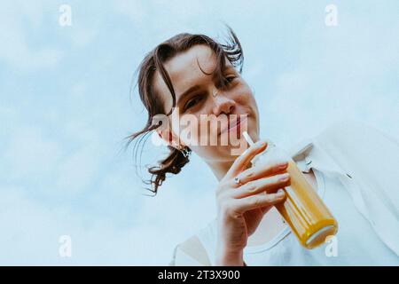 Niedrigwinkelporträt eines lächelnden jungen nicht-binären Menschen, der die Saftflasche gegen den Himmel hält Stockfoto