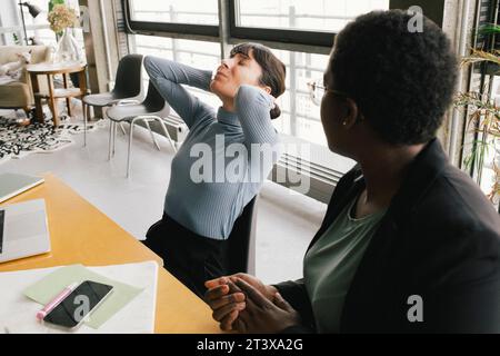 Geschäftsfrau, die neben überarbeiteter Kollegin sitzt und im Büro mit Schmerzen kämpft Stockfoto