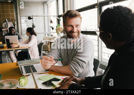 Lächelnder Geschäftsmann, der mit Geschäftsfrau spricht, während er am Schreibtisch im Büro sitzt Stockfoto