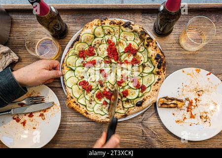 Männliche Hände schneiden eine frisch gebackene neapolitanische Zucchini-Pizza auf einem rustikalen Holztisch in einer traditionellen Pizzeria. Stockfoto