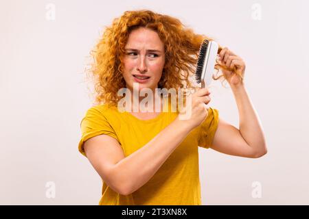 Wütende Ingwerfrau, die Haarbürste hält und ihr lockiges Haar kämmt. Stockfoto