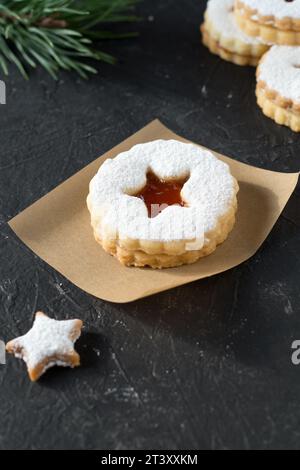 Leckere selbstgemachte Linzer-Kekse mit Marmelade und Zuckerpulver auf Papier zum Backen. Dunkler strukturierter Hintergrund mit Kiefernzweig. Stockfoto
